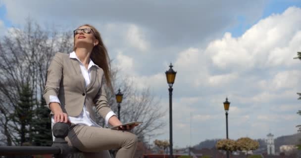 Peaceful pretty blonde businesswoman in good mood is enjoying the weather and nature, taking off her glasses and chatting via the tablet while sitting on the iron barrier. — Stock Video