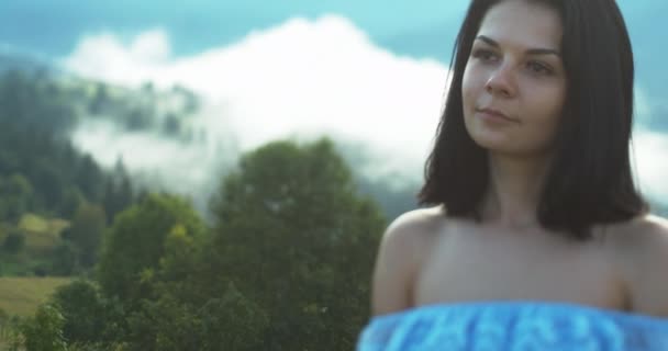 Potret luar ruangan berusia 20 tahun tersenyum berambut cokelat dengan gaun biru di latar belakang pegunungan ditutupi dengan kabut. — Stok Video