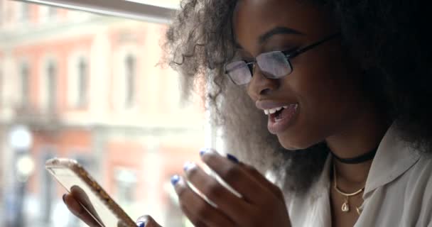 Retrato lateral de cerca de la sorprendida afroamericana con gafas sosteniendo el teléfono móvil y charlando cerca de la ventana. — Vídeo de stock