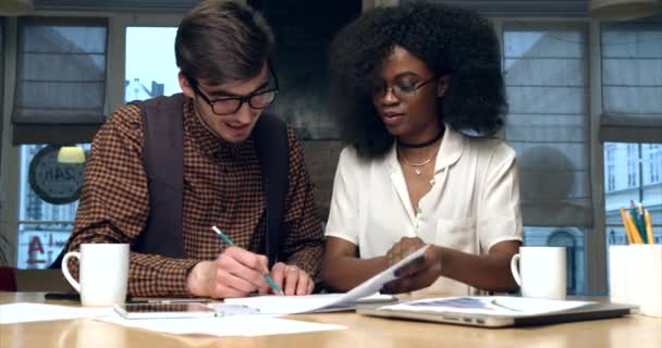 Csapatmunka két fiatal multikulturális üzletembertől szemüvegben, akik az irodai ábrákkal dolgoznak. Az ember leír valamit.. — Stock videók