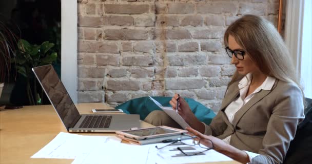 Närbild sida porträtt av den vackra blonda affärskvinna bär glasögon arbetar med dokument när du sitter på laptop på kontoret. — Stockvideo