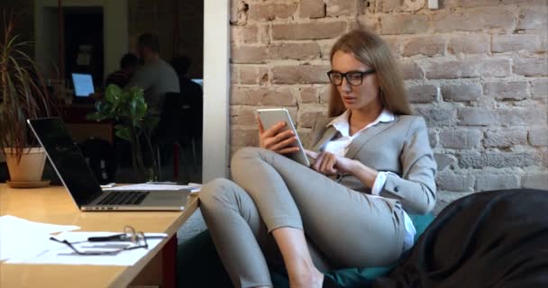 A gyönyörű, szemüveges üzletasszony a táblagépen dolgozik, miközben az irodában ül a kanapén.. — Stock videók