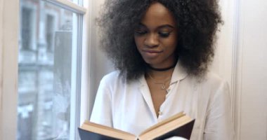 Çekici Afro-Amerikalı kadın pencere pervazında kitap okuyor. Yakın plan portre.