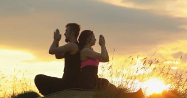 Erkek ve kadın arka arkaya lotus pozisyonunda oturuyorlar. Spor giyimli modern bir çift açık havada yoga egzersizleri yapıyor. 4K