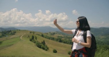 Güzel gülüşlü, doğal makyajlı ve sırt çantalı esmer turist çekici yeşil dağların arkasındaki cep telefonuyla selfie çekiyor. Yürüyüş konsepti. 4k görüntü.