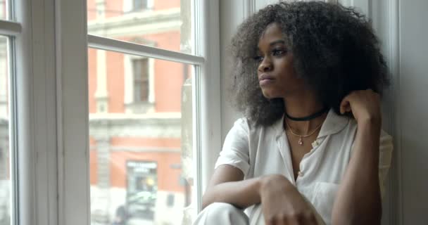 Den vackra unga upprörda afro-amerikanska kvinnan sitter på fönsterbrädan och tittar sorgligt nog genom fönstret.. — Stockvideo