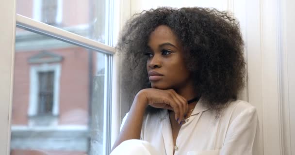 Portré a szomorú, gyönyörű afro-amerikai lányról, aki az ablakpárkányon ül, és boldogtalanul néz ki az ablakon.. — Stock videók