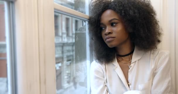 매력적 인 젊은아 프로 - 미국 여성 이 창가에 앉아, 기대고 슬프게 도 창을 통해 찾고 있습니다. 클로즈업 사진. 4k. — 비디오