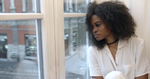 Portrait of the depressed charming young afro-american sitting on the window-sill and sadly looking on the street through the window. — Stock Video