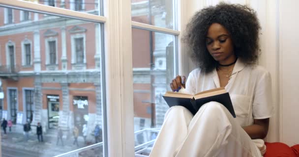 Joven mujer afroamericana atractiva está leyendo el libro mientras se relaja en el alféizar de la ventana. 4k. — Vídeo de stock