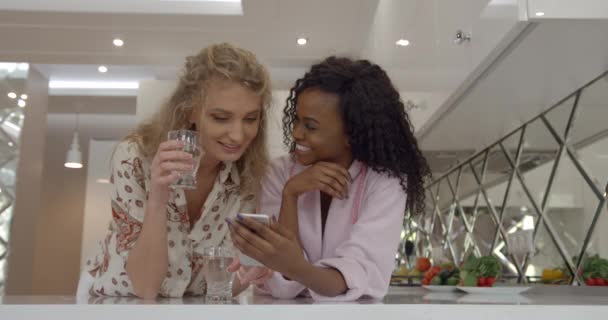 Mañana de dos hermosas africanas y encantadoras amigas caucásicas. Están hablando emocionalmente, sonriendo y viendo algo en el teléfono móvil en casa. Ubicación de la cocina moderna. — Vídeos de Stock