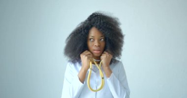 Doktor önlüklü, siyah kıvırcık saçlı çekici Afrikalı doktor beyaz arka planda steteskop kullanıyor..