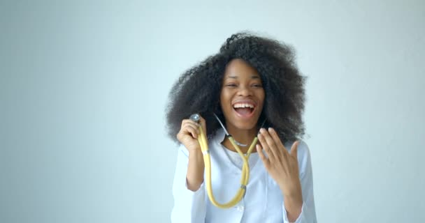 Šťastná krásná africká žena s přirozeným make-up v lékařském kabátu se baví při hraní se stetoskopem na bílém pozadí. Záběry 4k. — Stock video