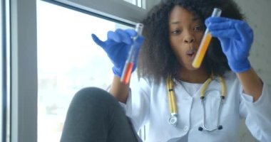 Gülümseyen Afrikalı kadın araştırmacı, kırmızı ve sarı kimyasal sıvıyı karıştırıyor ve pencere pervazında otururken test tüpünü sallıyor. 4k görüntü.