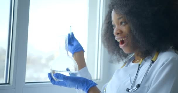 La sorprendida y feliz científica investigadora africana en bata de laboratorio mientras sostiene la pipeta y el frasco de fondo redondo con líquido cerca de la ventana. Imágenes de 4k. — Vídeos de Stock