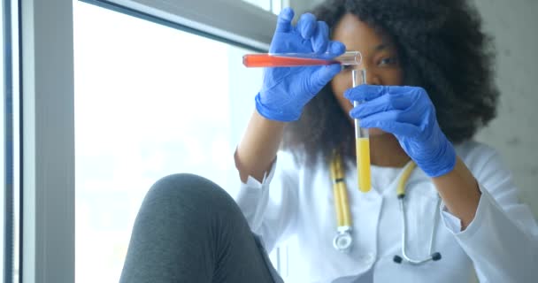 Una investigadora africana sonriente está llenando el líquido rojo de un tubo de ensayo a otro con líquido amarillo y agitándolo mientras está sentada en el alféizar de la ventana.. — Vídeos de Stock