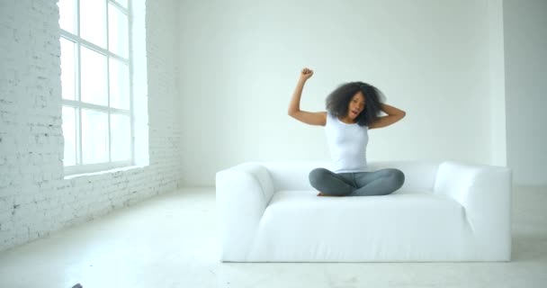 La giovane bella donna africana stanca con i capelli ricci in tessuto casual sbadiglia e si estende in posa di loto sul divano in camera bianca. 4k filmati. — Video Stock