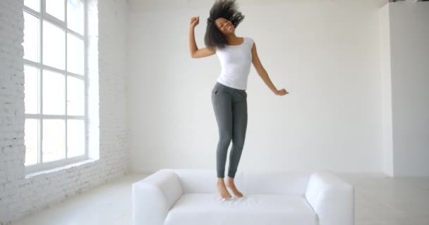 Çekici, mutlu, ev kıyafetleri içindeki Afrikalı kadın modern beyaz oturma odasındaki kanepede enerjik bir şekilde zıplarken eğleniyor. 4k görüntü. — Stok video