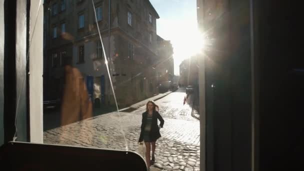 その美しい痩せっぽちの女の子が暗いカフェに入ってくる。ドアガラスを通して表示します。日当たりの良い通りを歩く. — ストック動画