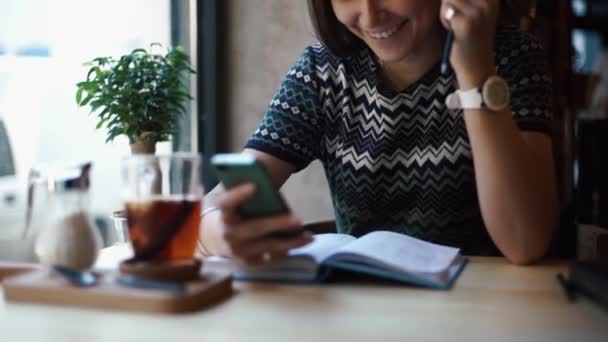 The young smiling girl is chatting, texting and browsing via the mobile phone while sitting in the cafe. She is enjoying her time with a cup of tea. — Stock Video