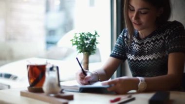 Not defterine yazan ve kafede çay içen gülümseyen genç kadının portresi..