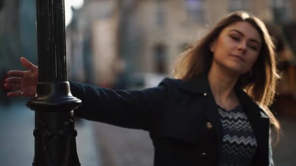 Mladá okouzlující dívka s přirozeným make-upem přichází k pouliční lampě a točí se kolem ní na slunné ulici. Koncept města. — Stock video