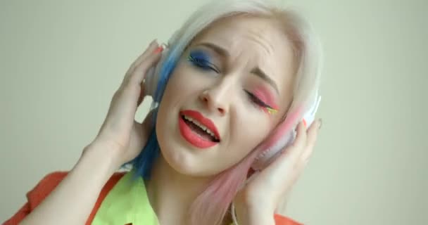 Hermosa mujer rubia con puntas de pelo rosa y azul y maquillaje colorido brillante es cantar y disfrutar de la música en los auriculares en el fondo blanco. — Vídeos de Stock