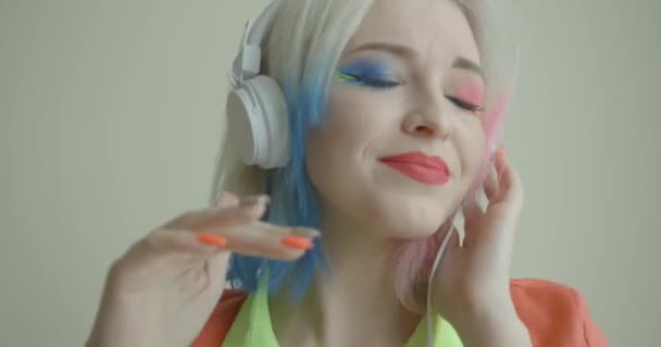 Känslomässigt porträtt av den attraktiva unga blonda kvinnan med blått och rosa hår slut och ljusa make-up lyssna på musik i hörlurar på vit bakgrund. Hon dansar och sjunger. — Stockvideo