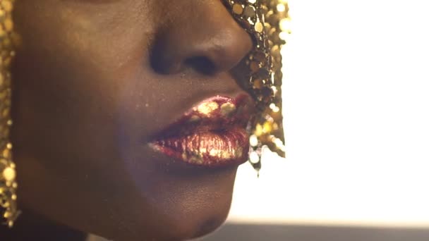 Labios rojos brillantes cubiertos con ollas de bronce dorado en la cara de una encantadora chica africana con joyas doradas en la cabeza. Primer plano . — Vídeos de Stock