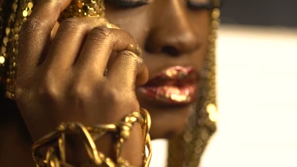美丽迷人的非洲女人的肖像，有着红润的嘴唇和金黄色的壶，青铜的眼睛和眼线，头上戴着金色的珠宝和手镯。她牵着她的手 — 图库视频影像