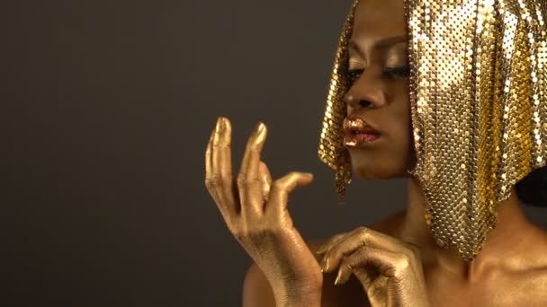 Attractive beautiful african woman with glossy red lips with golden pots, bronze eyeshadows and golden jewellery on head is opening and closing her golden fist and looking at it at the brown — Stock Video