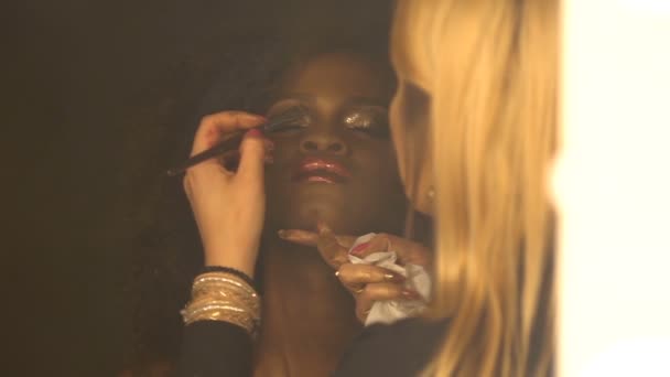 El artista de maquillaje prepara a la mujer africana joven hermoso con el pelo oscuro rizado a tiro. Ella está aplicando el brillo dorado de sus párpados. Reflexión en espejo. — Vídeos de Stock