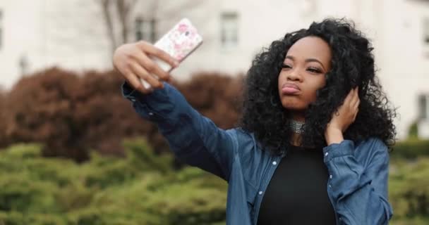 곱슬머리에 자연 화장을 한 젊고 아름다운 아프리카 소녀가 거리의 휴대폰을 통해 웃긴 얼굴로 셀카를 하고 있습니다.. — 비디오
