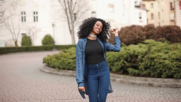 Het charmante slanke Afrikaanse meisje met natuurlijke make-up en mooie glimlach loopt vrolijk langs de straat terwijl ze haar donkere krullend haar aanraakt en schudt. — Stockvideo