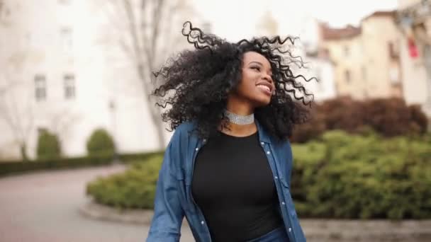 Close-up retrato da jovem menina africana atraente com maquiagem natural e sorriso bonito ativamente dançando e sacudindo seu cabelo encaracolado escuro ao ar livre. — Vídeo de Stock