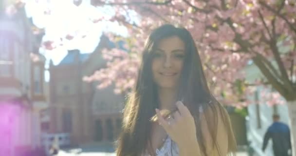 Jonge aantrekkelijke brunette met natuurlijke make-up en mooie glimlach speelt met haar haar en loopt langs de straat op de achtergrond van de bloeiende sakura. 4k-beelden. — Stockvideo