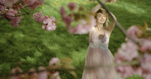 Uitzicht door bloeiende sakura boom op het mooie blonde meisje in roze jurk glimlachend terwijl u geniet van de natuur. 4k-beelden. — Stockvideo