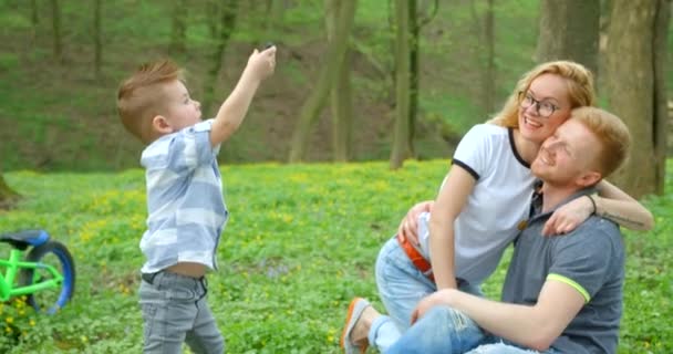 Pequeño niño lindo está tomando fotos de sus padres abrazos felices durante el picnic en el bosque verde. Imágenes de 4k. — Vídeos de Stock