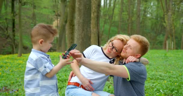 Pequeño niño lindo está tomando fotos de sus padres abrazos felices durante el picnic en el bosque verde. Imágenes de 4k. — Vídeos de Stock