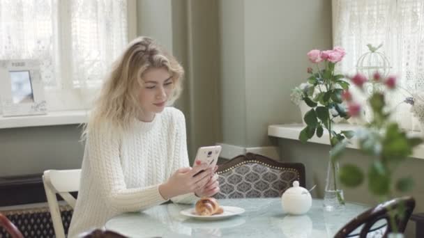 Piękna młoda blondynka z ładnym uśmiechem rozmawia, smsuje i przegląda przez telefon komórkowy podczas śniadania z rogalikiem. Materiał 4k. — Wideo stockowe