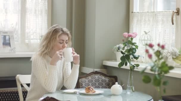 Güzel sarışın kız rahat bir kafede dinleniyor. O, kahvaltı sırasında kruvasanla kahve içiyor. 4k görüntü. — Stok video