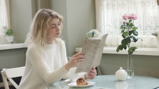 O retrato lateral da linda jovem loira atraente com maquiagem natural está lendo o menu enquanto se senta à mesa com croissant e buquê de rosas rosa em vaso. Localização do café. Filmagem 4k. — Vídeo de Stock