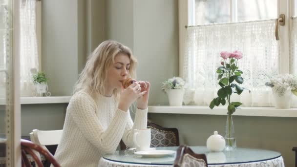 Doğal makyajlı güzel sarışın kız güzel bir kafede dinlenirken lezzetli kruvasanı kahveyle yiyor. O, pencereden bakıyor. 4k görüntü. — Stok video