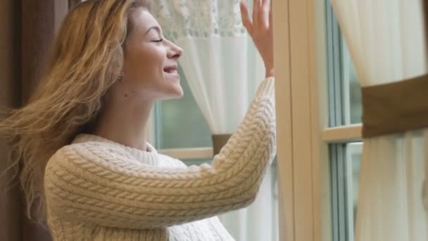 La giovane bella ragazza sorridente con il trucco naturale e muovendosi dai capelli del vento sta aprendo la finestra e salutando qualcuno, quindi chiudendolo. Ritratto ravvicinato. 4k filmati. — Video Stock