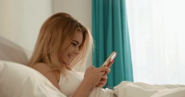 Sabah ve Dinlenme konsepti. Güzel gülüşü olan sevimli sarışın kız mutlu bir şekilde cep telefonunda sohbet ediyor ve yatakta geziniyor. Kulaklıkla müzik dinlemekten hoşlanıyor. 4k.