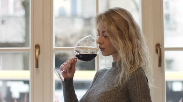 아름답고 예쁜 금발의 금발 이 커다란 유리에서 붉은 와인을 마시며 창문을 내다보고 있습니다. — 비디오