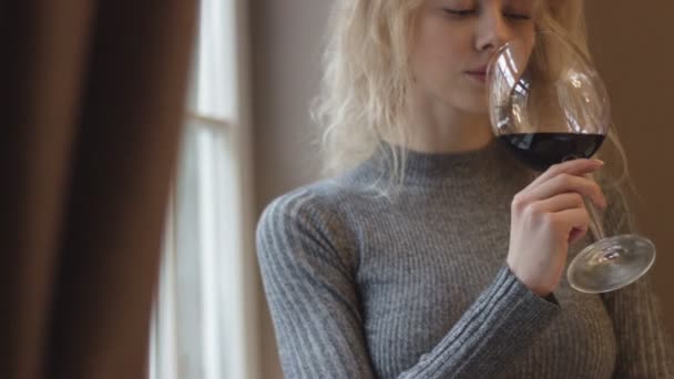 Όμορφη νεαρή ξανθιά κοπέλα με φυσικό μακιγιάζ πίνει κρασί απολαμβάνοντας τη θέα από το παράθυρο. 4k πλάνα. — Αρχείο Βίντεο