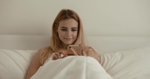 Porträt der glücklichen Frau, die lächelt, während sie im Bett plaudert und auf dem Handy surft. 4k. — Stockvideo