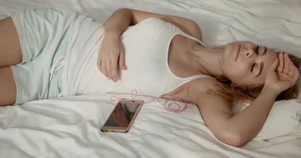 Beyaz pijamalı güzel genç kız, gözleri kapalı yatakta dinlenirken kulaklıkla müzik dinliyor. 4k görüntü. — Stok video