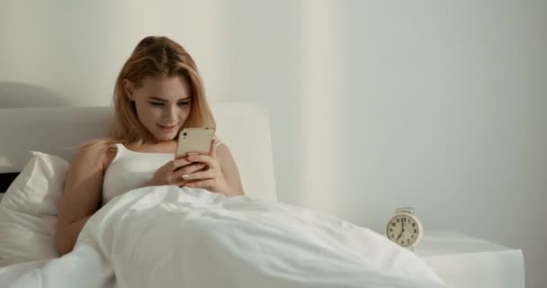 Την ώρα του ύπνου. Πορτρέτο του πανέμορφου φρέσκο ξανθό κορίτσι με ελκυστικό χαμόγελο ευτυχώς κουβέντα και περιήγηση στο κινητό τηλέφωνο, ενώ βρίσκεται στο κρεβάτι. 4ια. — Αρχείο Βίντεο